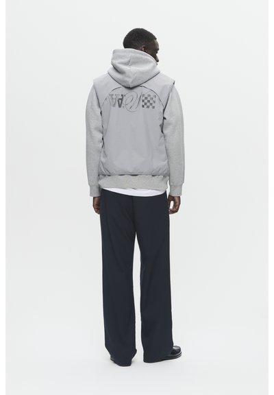 Veste bomber grise avec capuche et manches grises contrastantes, arborant un logo à carreaux au dos, associée à un pantalon large bleu marine.