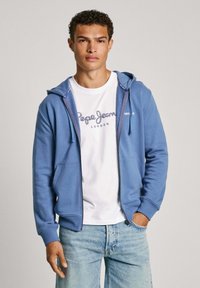 Zils slēdzams kapuci džemperis, izgatavots no mīkstas auduma, ar kangaroo kabatu, uzvilkts virs baltas T-kreklu ar zilu logotipu, kombinēts ar gaiši zilām džinsām.