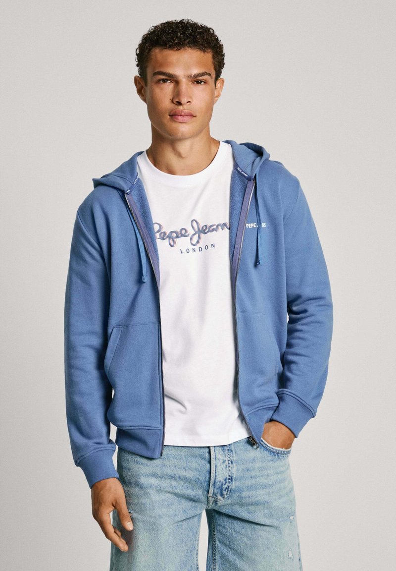 Zils slēdzams kapuci džemperis, izgatavots no mīkstas auduma, ar kangaroo kabatu, uzvilkts virs baltas T-kreklu ar zilu logotipu, kombinēts ar gaiši zilām džinsām.