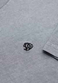 Grå bomuld t-shirt med rund halsudskæring, der har en lille sort broderet logo på brystet og korte ærmer.