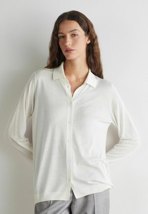 Frau mit langen lockigen Haaren, die eine weiße, geknöpfte Strickjacke und graue Hose trägt, steht mit den Händen in den Hüften vor einer schlichten hellen Wand.