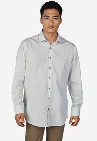 Mann mit kurzem schwarzen Haar, der ein hellgraues Hemd mit kleinen Punkten und braune Hose trägt, vor einem weißen Hintergrund stehend.