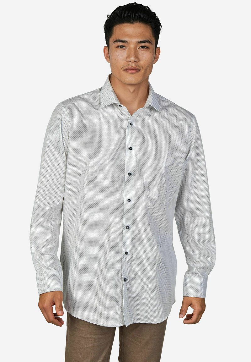 Mann mit kurzem schwarzen Haar, der ein hellgraues Hemd mit kleinen Punkten und braune Hose trägt, vor einem weißen Hintergrund stehend.