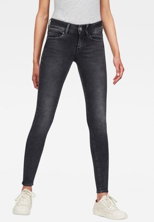 Femme portant un jean skinny noir assorti de baskets blanches et d'un haut gris sans manches, debout les jambes écartées sur un fond blanc.