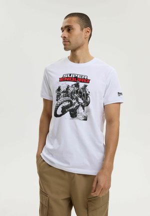 Mężczyzna w białej koszulce z grafiką wyścigów motocyklowych i napisem "SUPER ENDURO", ubrany w beżowe spodnie cargo, stoi na jednolitym tle.