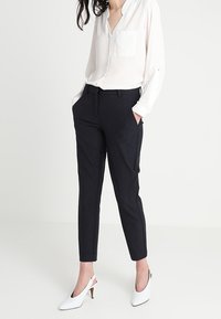 White button-up shirt with a pocket, paired with dark tapered trousers, featuring slanted pockets and white heels with a slender heel.