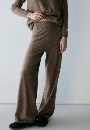 Personne portant un pantalon en maille marron ample et un haut à manches longues assorti, soulevant légèrement le haut pour révéler la taille, debout sur un sol clair.
