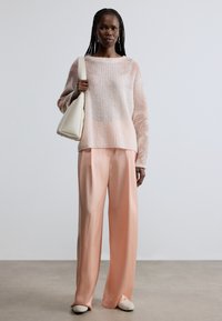 Hellrosa gestrickter Pullover mit weiten Ärmeln, kombiniert mit locker sitzender lachsfarbener Satinhose. Das Modell hält eine cremefarbene Handtasche.