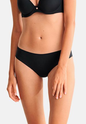 Femme portant un soutien-gorge noir et un shorty noir sans coutures, debout les bras le long du corps, teint clair, fond uni.