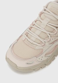 Light beige sneaker with mesh and leather, featuring pink accents, thick sole, and prominent lacing system. Round toe and textured surface.