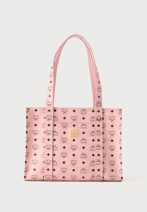 Różowa torba typu tote MCM z czarnym wzorem logo, złotą plakietką oraz dwoma długimi rączkami, pokazana na gładkim jasnym tle.