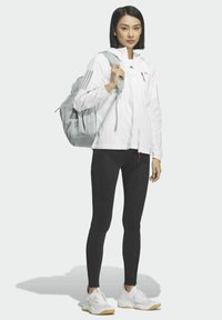 Giacca bianca con zip e tre strisce nere, leggings neri e scarpe da ginnastica bianche. Zaino grigio indossato su una spalla, design minimalista.