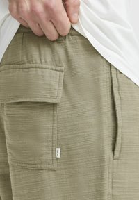 Olivengrüne Cargo-Shorts aus strukturiertem Stoff, mit einer Seitentasche, subtilen Nähten und einem kleinen Emblem auf der Tasche.