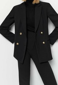 Blazer noir à double boutonnage avec des boutons dorés, associé à un col roulé noir, présentant une coupe ajustée et une texture de tissu lisse.