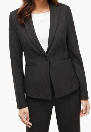 Mujer con blazer entallado gris oscuro y pantalones a juego sobre una camiseta blanca, con dos collares plateados en capas.