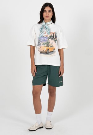 Donna che indossa una T-shirt bianca con stampa di un'auto d'epoca, pantaloncini verdi, foulard azzurro chiaro con motivi, calzini bianchi e mocassini beige.