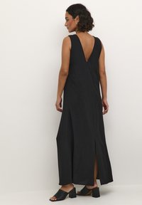 Black sleeveless maxi dress with a deep V-back, side slits, and smooth fabric. Paired with black open-toe chunky heel mules.