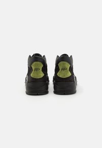 Pár černých tenisek s olivově zelenými semišovými záplatami na patách a černým logem, viděno zezadu na bílém pozadí.