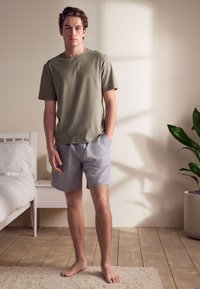 T-shirt vert olive à manches courtes avec une coupe décontractée, associée à un short gris clair. Texture de tissu douce, debout sur un sol en bois.