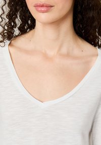 Balta V formos palaidinė laisvu siluetu, pasižyminti švelniu, lengvu tekstūru ir subtiliu horizontaliu raštu. Atidengia kaklo sritį.