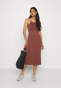 Vestido midi castanho, justo, com alças finas, feito de tecido suave. A modelo segura uma mala tote preta texturizada e usa ténis claros.