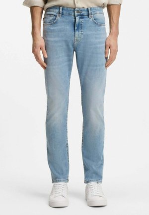 Man draagt lichtblauwe slim-fit jeans en witte sneakers, staat met de handen ontspannen langs het lichaam tegen een effen achtergrond.