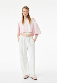 Top corto rosa con letras blancas, combinado con pantalones de chándal blancos holgados y zapatillas rosas tipo slip-on. Tejido suave, ajuste relajado y minimalismo.