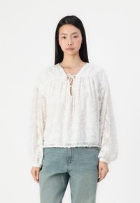 Weiße, transparente Bluse mit floraler Stickerei, V-Ausschnitt und Bindedetail am Ausschnitt. Lockere, lange Ärmel und ein verkürzter Saum. Kombiniert mit Jeans.