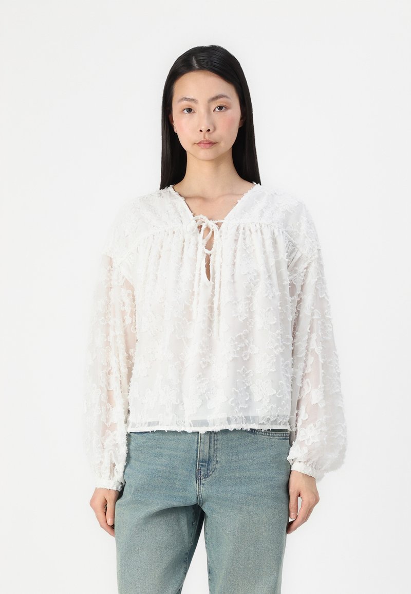 Weiße, transparente Bluse mit floraler Stickerei, V-Ausschnitt und Bindedetail am Ausschnitt. Lockere, lange Ärmel und ein verkürzter Saum. Kombiniert mit Jeans.