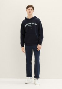 Felpa navy con scritta "Denim & Tailor", dotata di tasca frontale e cappuccio con lacci, abbinata a pantaloni navy e sneakers bianche.