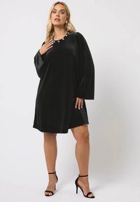 Schwarzes Samtkleid mit langen Ärmeln, rundem Ausschnitt und floralen Akzenten. A-Linien-Silhouette, knielang mit Seitenschlitzen, kombiniert mit schwarzen Absätzen.