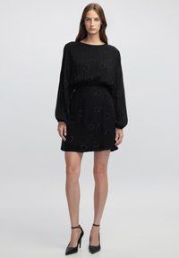 Schwarzes Kleid mit langen Ärmeln und rundem Ausschnitt, verziert mit kreisförmigen Perlenmustern. Glattes Material und kniebedeckende Länge.