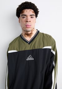 Adidas långärmad jacka med v-ringning i olivgrön och svart, med vita detaljer och logotyp på bröstet; slät textur, avslappnad passform.