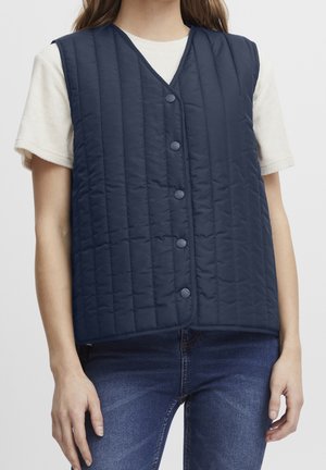 Vrouw draagt een marineblauw gewatteerd vest met knopen over een wit shirt met korte mouwen en een blauwe spijkerbroek, afgebeeld van nek tot heupen.