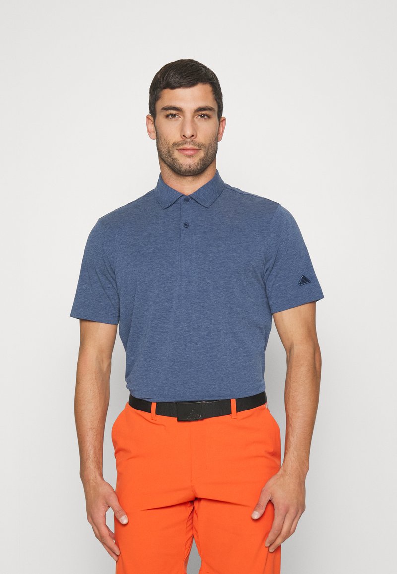 Polo de manga corta azul oscuro con cuello, tres botones y un pequeño logotipo en el hombro izquierdo. Combinado con pantalones naranja brillante.