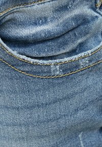 Lichtblauwe denimstof met zichtbare stikseldetails, met een contour van een zak en lichte slijtage, die een gladde textuur toont.