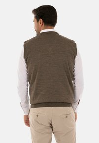 Gilet di maglia marrone con scollatura profonda e senza maniche, indossato sopra una camicia bianca a maniche lunghe, abbinato a pantaloni beige.