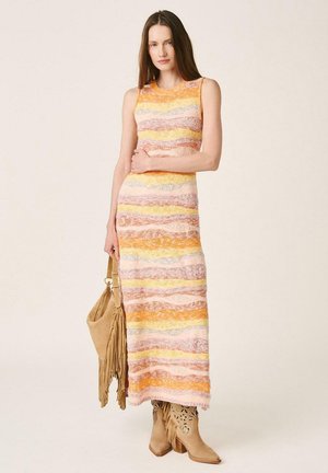 Femme portant une robe maxi sans manches à rayures multicolores, tenant un sac à main beige à franges, et portant des bottines beige à franges.