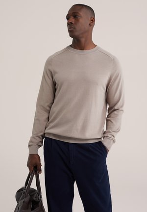 Mann trägt einen beigen Pullover und dunkle Hose, hält eine schwarze Ledertasche und schaut seitlich vor hellem, einfarbigem Hintergrund.