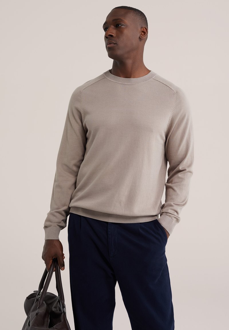 Homme portant un pull beige et un pantalon sombre, tenant un sac en cuir noir, regardant sur le côté devant un fond clair uni.