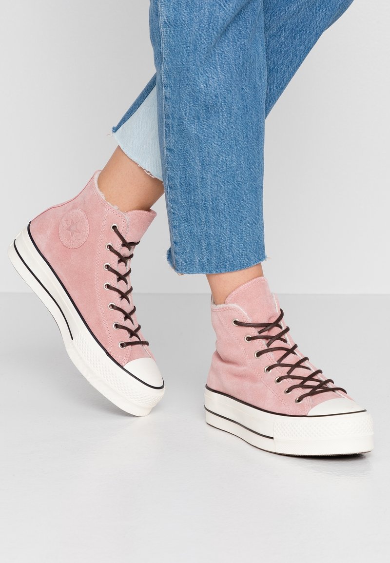Personne portant un jean bleu et des baskets montantes roses avec des semelles blanches et des lacets noirs, debout sur un sol gris clair.