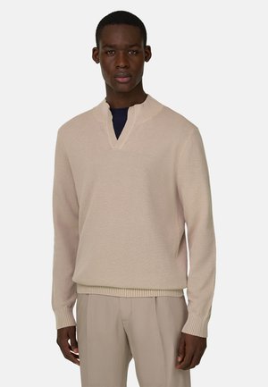 Man draagt een beige getextureerde trui met V-hals en bijpassende beige broek, staand tegen een effen lichte achtergrond.