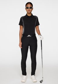 Kobieta w czarnym stroju golfowym, w białej rękawiczce i okularach przeciwsłonecznych, trzymająca kij golfowy, stojąca na tle jednorodnego białego tła.