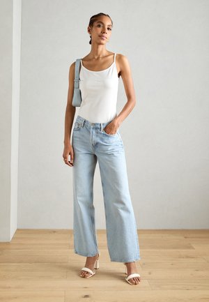 Débardeur blanc ajusté, jeans bleu clair à jambes larges et sandales à talons blancs. La tenue présente un design minimaliste avec une silhouette décontractée.