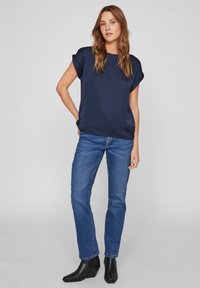 Marineblå satinbluse med korte opbrettede ærmer, kombinert med blå rette jeans og svarte hælhøye ankelstøvler. Enkel struktur, uten mønstre.