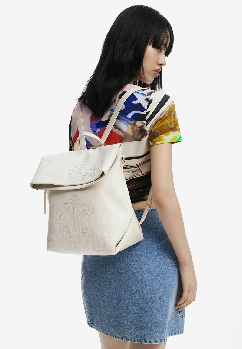 Zaino in pelle bianca con design floreale impresso, dotato di spallacci regolabili e patta pieghevole, indossato sopra una camicia a fantasia colorata.