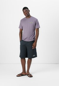 Lilla bomulls T-skjorte med liten grafikk, kombinert med svarte shorts med hvit broderi, og svarte flip-flops på en ensfarget bakgrunn.