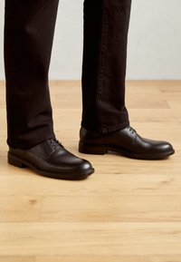 Chaussures noires en cuir à lacets avec une finition lisse, bout arrondi et talon bas, associées à un pantalon sombre sur un sol en bois.