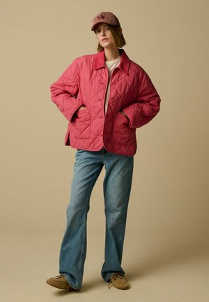 Femme portant une veste matelassée rose, un jean bleu clair, des chaussures marron et une casquette en velours côtelé rose, debout devant un fond beige.