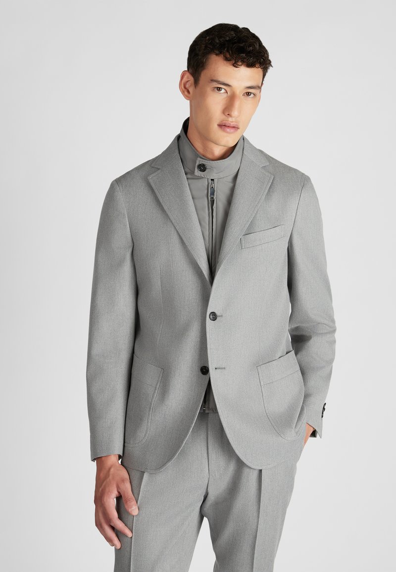 Completo grigio su misura con colletto a revers, due tasche frontali e chiusura con bottoni, indossato sopra una giacca grigia con zip.